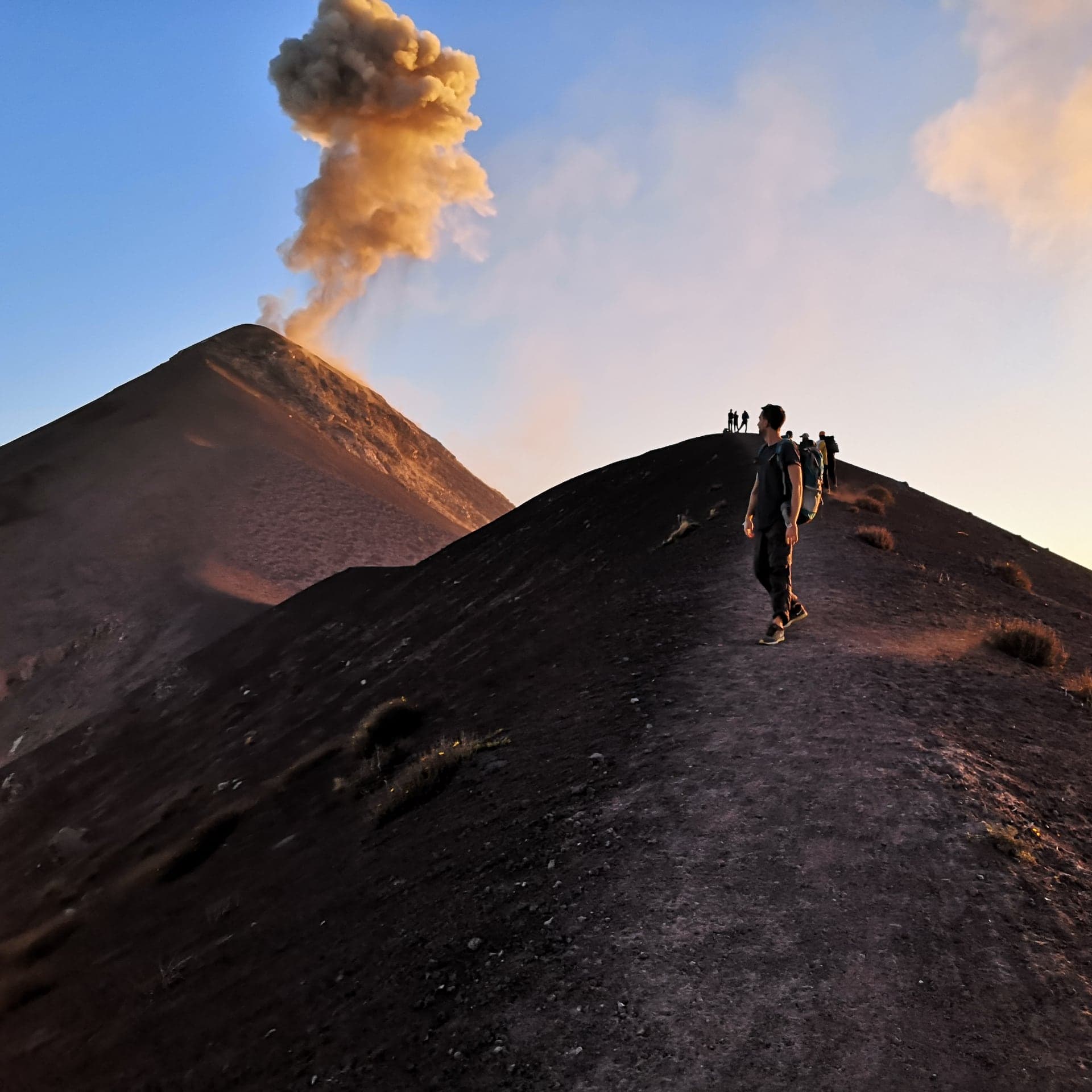 A walker on a volcanic ridge facing an erupting volcano at sunrise